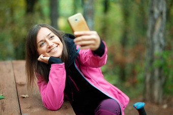 徒步旅行者女孩休息板凳上森林背包客粉红色的夹克采取自拍智能手机