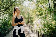 水平拍摄活跃的运动女人智能手机检查结果慢跑穿着活跃的穿享受温暖的阳光明媚的一天听音乐耳机健康的生活方式概念
