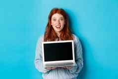 兴奋红色头发的人女自由职业者显示移动PC屏幕盯着相机惊讶站电脑蓝色的背景