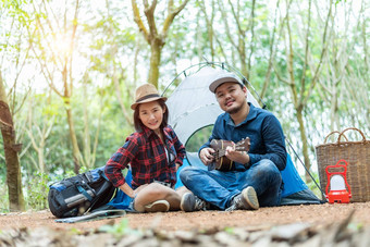亚洲夫妇野营森林男人。玩尤克里里琴女人前面野营帐篷灯篮子背包元素人户外生活方式概念冒险旅行主题