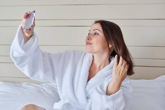美丽的成熟的女人采取自拍智能手机坐着白色浴袍床上