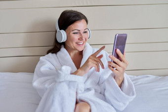 快乐成熟的女人<strong>白色浴袍</strong>耳机智能手机坐着床上