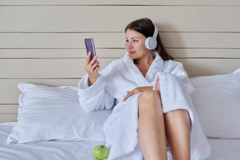 快乐成熟的女人白色浴袍耳机智能手机坐着床上