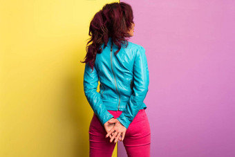 美丽的年轻的女人紫色的头发穿蓝色的皮革夹克品红色的牛仔裤站构成孤立的彩色的背景