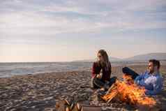 年轻的夫妇坐着海滩篝火喝啤酒
