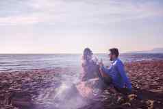 年轻的夫妇坐着海滩篝火喝啤酒