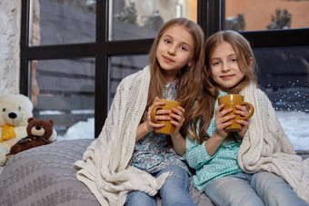 图像可爱的姐妹坐着窗台上覆盖毯子喝温暖的茶