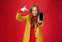 选择电话后悔肖像友善的有吸引力的自信红色头发的人魅力女朋友黄色的外套显示智能手机指出移动微笑广泛的相机