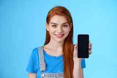 特写镜头有吸引力的快乐的红色头发的人年轻的女商店助手建议客户买智能手机显示电话显示促进应用程序互联网应用程序微笑相机站蓝色的背景