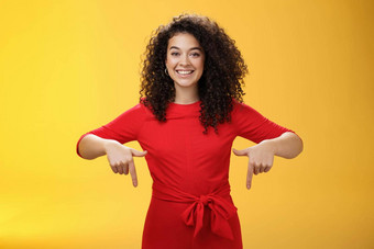 工作室拍摄愉快的微笑年轻的卷发女人红色的衣服指出指数手指促进太棒了事件微笑广泛的建议买黄色的墙