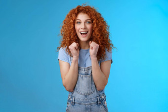 快乐的乐观幸运的红色头发的人卷发有吸引力的女人欢呼握紧的拳头快乐微笑广泛的欢呼看游戏支持鼓励动机蓝色的背景