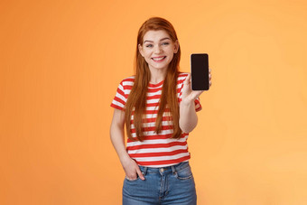 快乐的好看的红色头发的人女人<strong>介绍</strong>社会媒体<strong>功能产品</strong>持有智能手机显示相机手机显示促进设备应用程序微笑广泛的橙色背景