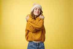乐观的迷人的快乐的年轻的女人感觉幸运的快乐拥抱温暖的舒适的毛衣倾斜头肩膀关闭眼睛微笑广泛的拥抱身体黄色的背景