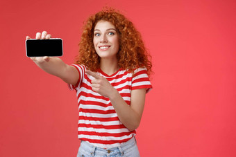 兴奋快乐好看的红色头发的人卷曲的女显示水平智能手机屏幕指出显示小工具微笑很高兴骄傲击败朋友分数游戏站红色的背景
