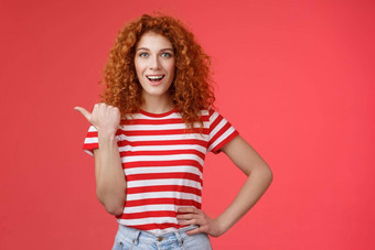 时髦的好看的感情的快乐微笑红色头发的人欧洲女卷曲的发型指出拇指左露齿而笑自信的厚颜无耻的持有手腰导演促销广告提供红色的背景