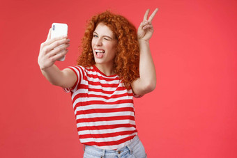 时髦的时尚好玩的好看的红色头发的人大胆的卷曲的女人显示和平胜利动物耳朵手势眨眼智能手机显示记录视频博客采取自拍太棒了电话相机红色的背景