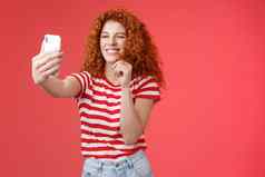 时髦的好看的时尚的有魅力的红色头发的人女卷曲的发型眨眼厚颜无耻的表达式使轻浮古怪的脸持有智能手机采取自拍记录视频消息玩有趣的面部过滤器