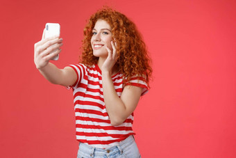 受欢迎的快乐的好看的时尚的女博主红色头发的人卷曲的发型感觉漂亮的自我接纳采取自拍提高了手臂持有智能手机摆姿势愚蠢的可爱的电话相机红色的背景