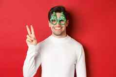 图像英俊的男人。白色毛衣聚会，派对眼镜显示和平标志微笑祝快乐圣诞节站红色的背景