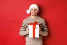 图像英俊的微笑男人。圣诞老人他冬天毛衣持有现在给圣诞节礼物祝快乐假期站红色的背景