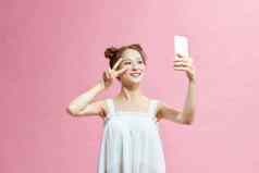可爱的好玩的年轻的女人采取自拍移动电话粉红色的背景
