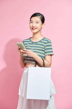 时尚的美丽的年轻的亚洲女人携带色彩斑斓的袋购物在线移动电话孤立的粉红色的背景