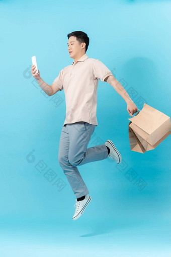 兴奋年轻的英俊的亚洲男人。持有移动电话购物袋跳光蓝色的工作室孤立的背景