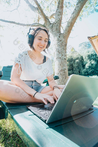 年轻的女人坐着花园微笑研究大学享受户外娱乐听力音乐移动PC房子研究人生活方式概念打字电脑