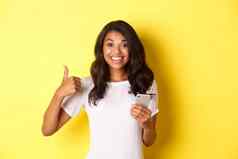 肖像年轻的好看的非洲美国女人显示竖起大拇指移动电话应用程序微笑很高兴站黄色的背景