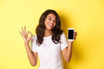 肖像可爱的非裔美国人女孩微笑很高兴显示标志移动电话屏幕推荐应用程序促销站黄色的背景