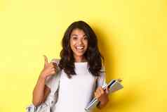 图像快乐的非裔美国人女孩学生持有笔记本电脑类背包显示竖起大拇指研究站黄色的背景