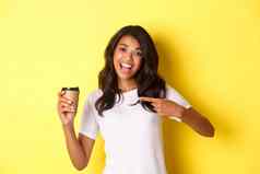 图像快乐的非裔美国人女人推荐好咖啡咖啡馆指出杯站黄色的背景