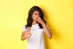 年轻的非裔美国人女人白色t恤关闭鼻子恶心保持不喜欢坏气味站黄色的背景