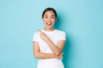 肖像兴奋快乐美丽的亚洲女孩乐观的会说话的广告指出手指上左角落里显示促销提供微笑惊讶蓝色的背景