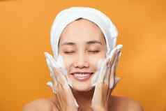 快乐年轻的亚洲女人应用脸奶油穿毛巾触碰脸孤立的橙色背景