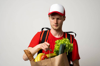 杂货店交付快递男人。红色的帽t恤热袋肩膀持有纸袋食物白色工作室背景食物交付服务在线购物