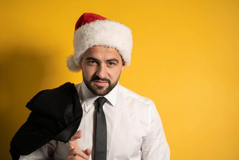 英俊的年轻的男人。穿圣诞老人他圣诞节季节准备好了祝贺同事英俊的微笑有胡子的男人。圣诞老人他孤立的黄色的背景