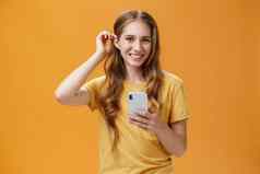 半身照拍摄可爱的友善的不错的年轻的女孩波浪发型把头发海滩耳朵持有智能手机微笑广泛的快乐的表达式相机橙色背景