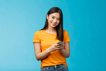 快乐的友好的现代年轻的亚洲女孩学生消息传递朋友持有智能手机发短信追随者互联网应用程序应用程序站满意微笑相机蓝色的背景