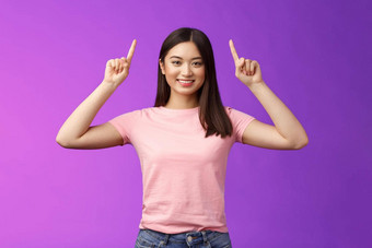 可爱的自信的黑头发的亚洲女人穿粉红色的t恤提高手显示促销指出手指咧着嘴笑邀请链接试演产品给方向站紫色的背景
