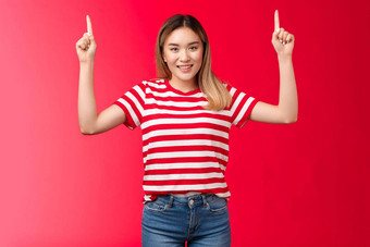 嘿快乐的好看的年轻的亚洲女孩指出手显示促销活动微笑快乐的女模型提出好的地方挂广告站红色的背景