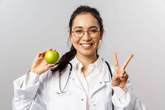 肖像热情的年轻的快乐微笑女医生白色外套显示和平标志绿色苹果人保持健康的吃水果维生素灰色背景