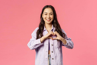 肖像可爱的有吸引力的亚洲女孩牛仔布夹克微笑显示心标志承认爱浪漫的表达同情充满激情的感觉下降粉红色的背景