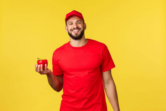 在线交付购物食物订单概念英俊的快乐的快递红色的统一的帽t恤持有新鲜的胡椒交付食品杂货商店客户首页