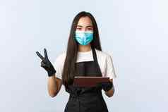 科维德在线订单小咖啡商店业务防止病毒概念可爱的友好的亚洲咖啡师女咖啡馆工人医疗面具手套显示和平标志持有数字平板电脑