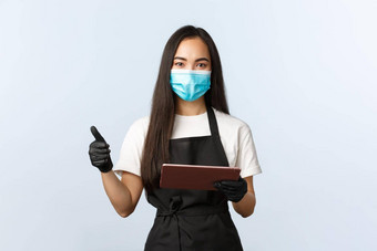 科维德在线订单小咖啡商店业务防止病毒概念微笑可爱的亚洲女咖啡师围裙医疗面具手套显示翘拇指订单数字平板电脑
