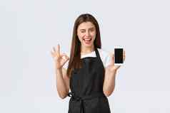 杂货店商店员工小业务咖啡商店概念很高兴好看的女孩咖啡师黑色的围裙显示标志移动电话显示推荐很酷的应用程序