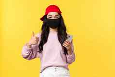 科维德社会距离生活方式防止病毒传播概念微笑快乐的亚洲女孩红色的帽脸面具保护健康冠状病毒使竖起大拇指移动电话