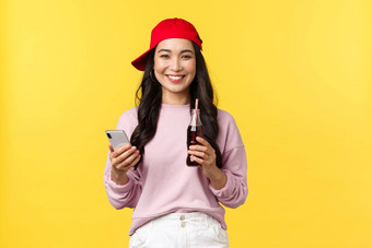 人情绪饮料夏天休闲概念年轻的十几岁的朝鲜文女孩红色的帽消息传递智能手机喝苏打水饮料站黄色的背景很高兴