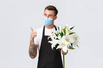 冠状病毒社会距离业务科维德流感大流行概念可爱的友好的花店推销员花显示显示竖起大拇指微笑持有花束百合穿医疗面具